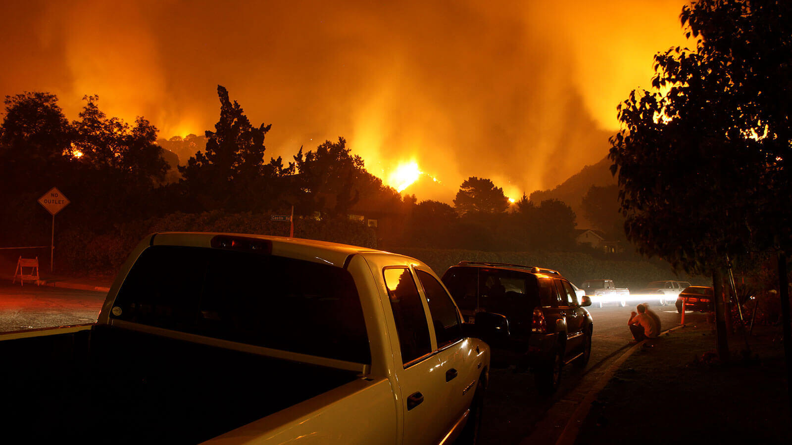 Cars lining up on a street watching a forest fire behind the tree line.