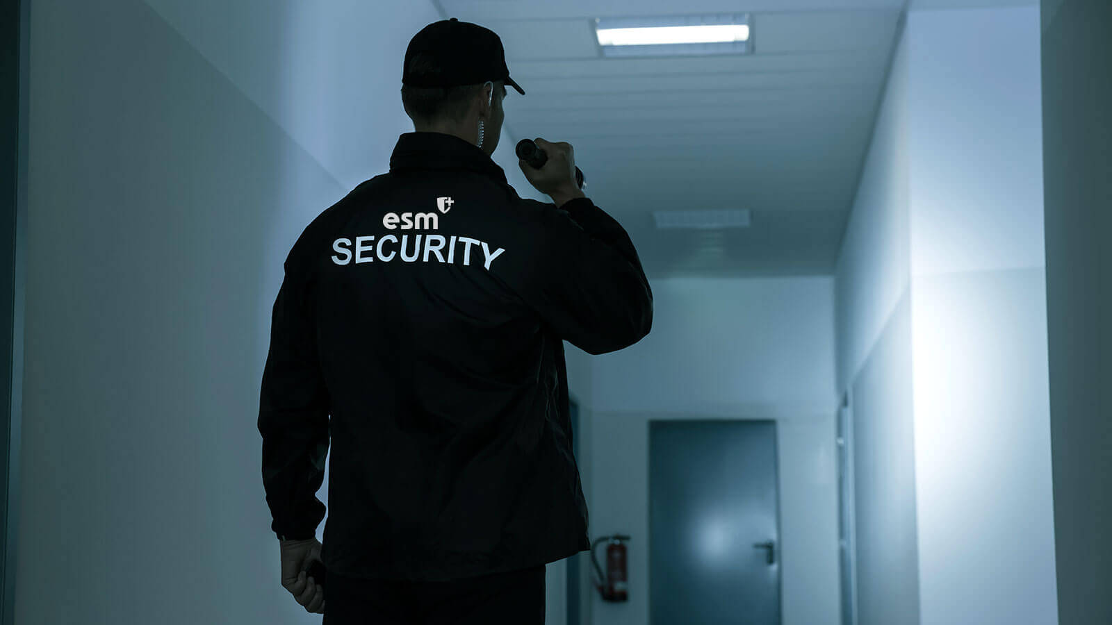 Security guard checking a hallway with a flashlight in security jacket and hat.